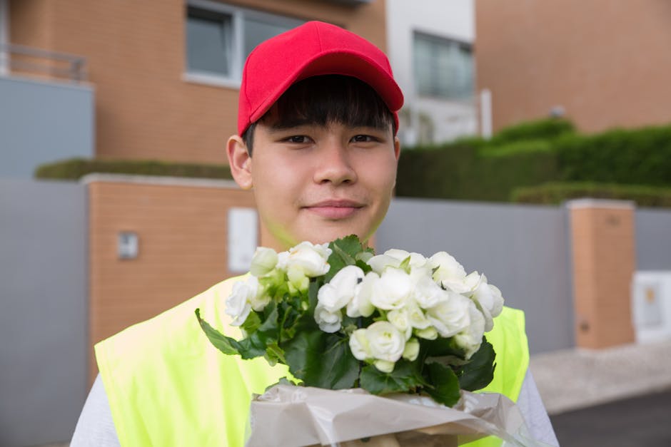 A young delivery person wearing a red cap and neon yellow vest is standing outdoors in front of modern residential buildings, holding a bouquet of fresh white flowers wrapped in white paper. The bouqu