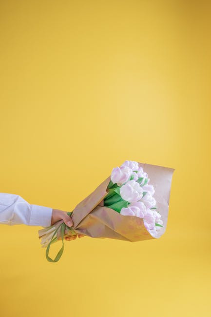 A person dressed in a white shirt is holding a bouquet of fresh tulips wrapped in light brown paper with a green ribbon, positioned in front of a solid yellow background. The tulips have pale pink ope