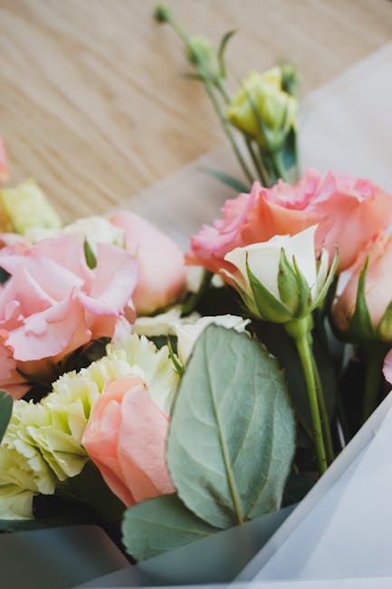 A close-up of a floral arrangement featuring soft pink and cream lisianthus flowers with ruffled petals, complemented by fresh green leaves, placed on a wooden surface. The bouquet is wrapped in light