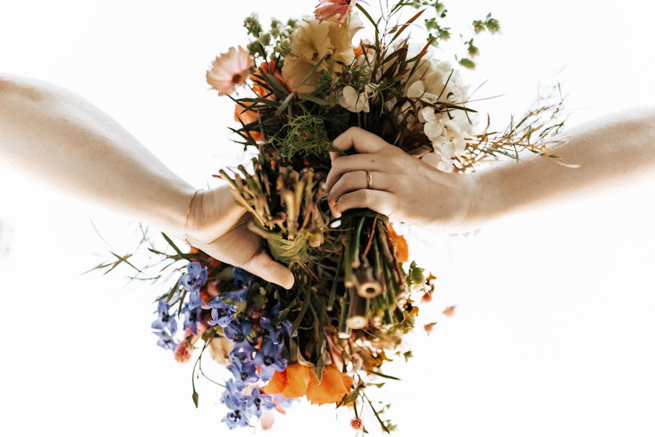Two hands, one with a gold ring, holding a vibrant bouquet of mixed fresh flowers including orange poppies, light pink roses, purple delphiniums, and white hydrangeas, wrapped in natural brown paper t
