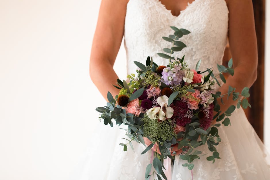 A bride in a white lace wedding dress holds a large, vibrant bouquet featuring a mix of flowers including white orchids, pink and purple chrysanthemums, burgundy and mauve roses, purple hydrangeas, an