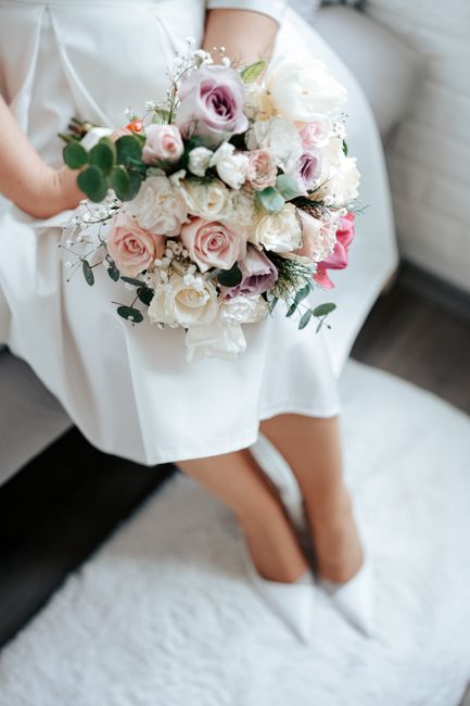 A person dressed in a white garment is seated on a soft surface, holding a large bouquet of fresh flowers close to their lap. The bouquet features a variety of blossoms including pale pink and lavende