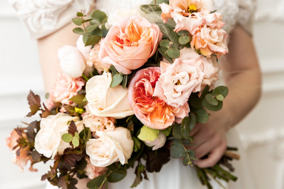 A person holding a bouquet of fresh flowers featuring peach and cream roses, pink lisianthus, and eucalyptus leaves, wrapped in white tissue paper. The bouquet is positioned in front of the person's t