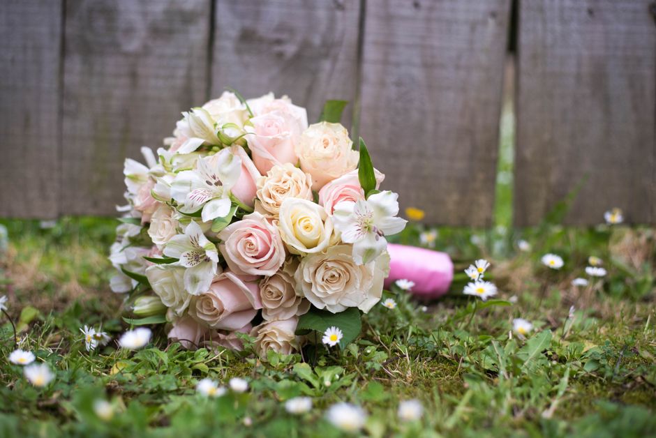 A pastel-colored floral bouquet resting on grass in an outdoor setting, featuring pale pink roses, creamy white roses, and white lilies with light pink streaks, all freshly arranged with green foliage