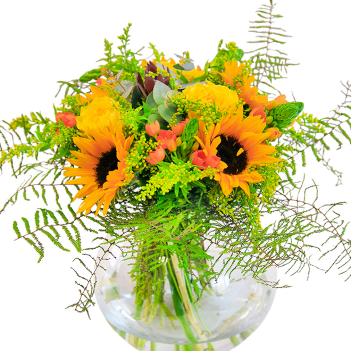 Sunlit exuberance spills from this lush bouquet, expertly arranged in a round glass vase and radiating the joyful spirit of Queensbury. Broad faces of sunflowers bloom with confidence, their golden-yellow petals fanning out in cheerful circles around inky brown centers, suggesting an endless supply of summer warmth like the open spaces of Canon Lane. Alongside, tightly packed yellow garden roses bloom in sophisticated spirals, each petal soft and velvety, contributing classic luxury and refinement. Glossy red Hypericum berries dot the composition, bringing a sparkling zest and contrast against the dominant yellows and verdant foliage. Cascading ferns, as feathery as those in local autumn woodlands near Queensbury, provide depth and a gentle sway. Airy goldenrod or solidago sprigs add a delicate hint of wildness, their minuscule yellow flowers echoing sunlight filtering through leafy branches. Subtle eucalyptus leaves, tinged with blue-green, peek out to create a fresh, invigorating touch. The transparent round vase reveals the bouquet's structural artistry-the crisscross of green stems immersed in clear water, fostering a serene and inviting presentation. Every element of this arrangement feels thoughtfully chosen, its scent a delicate blend of floral brightness and leafy freshness. It's a perfect showpiece for celebrations, anniversaries, or as a loving expression to uplift someone in the heart of Queensbury, crafted to bring the outdoors in and flood any room with pure, radiant joy.